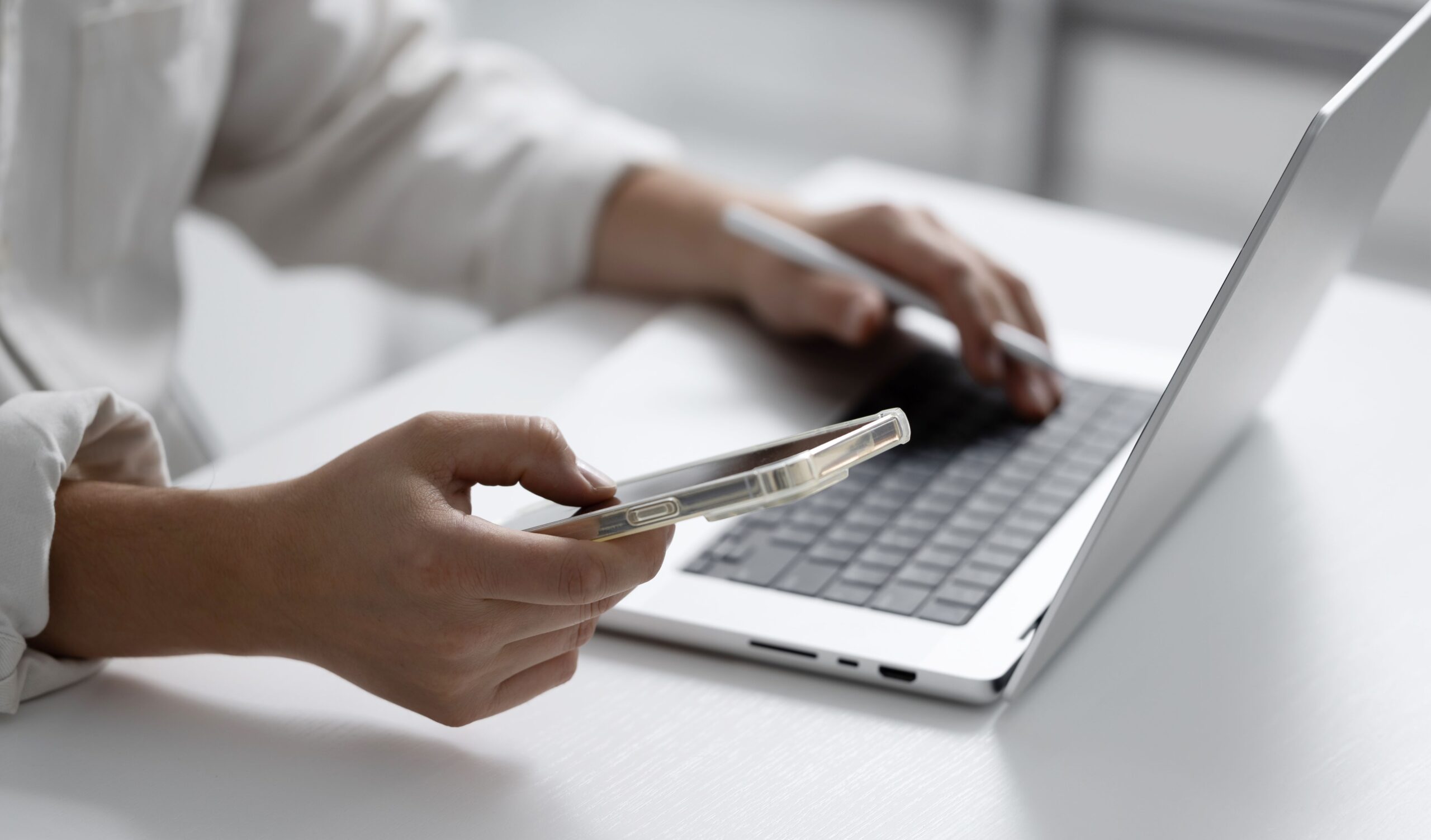 A woman holds an iPhone and works on her computer at a desk.